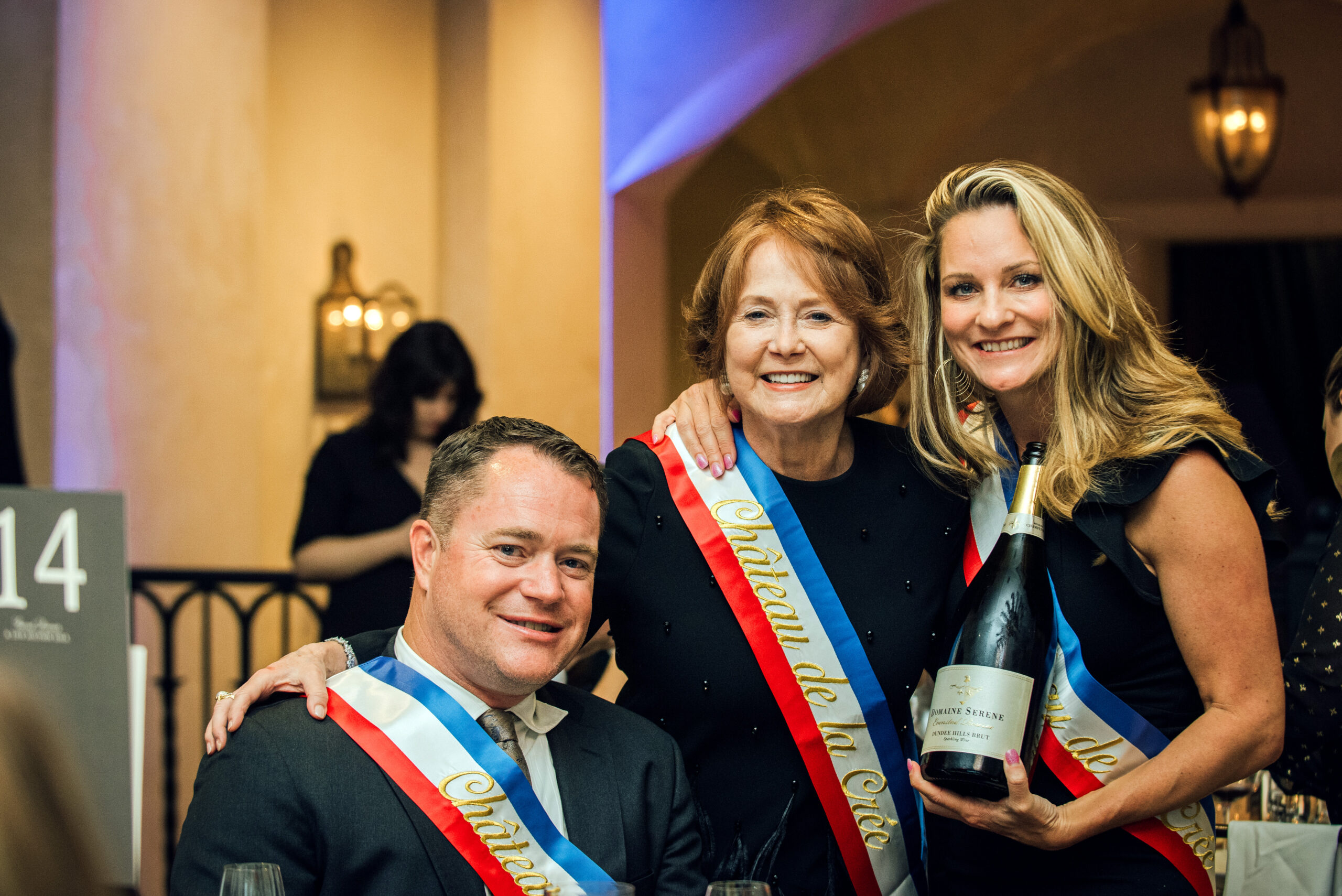 People wearing celebratory sashes with a bottle of wine